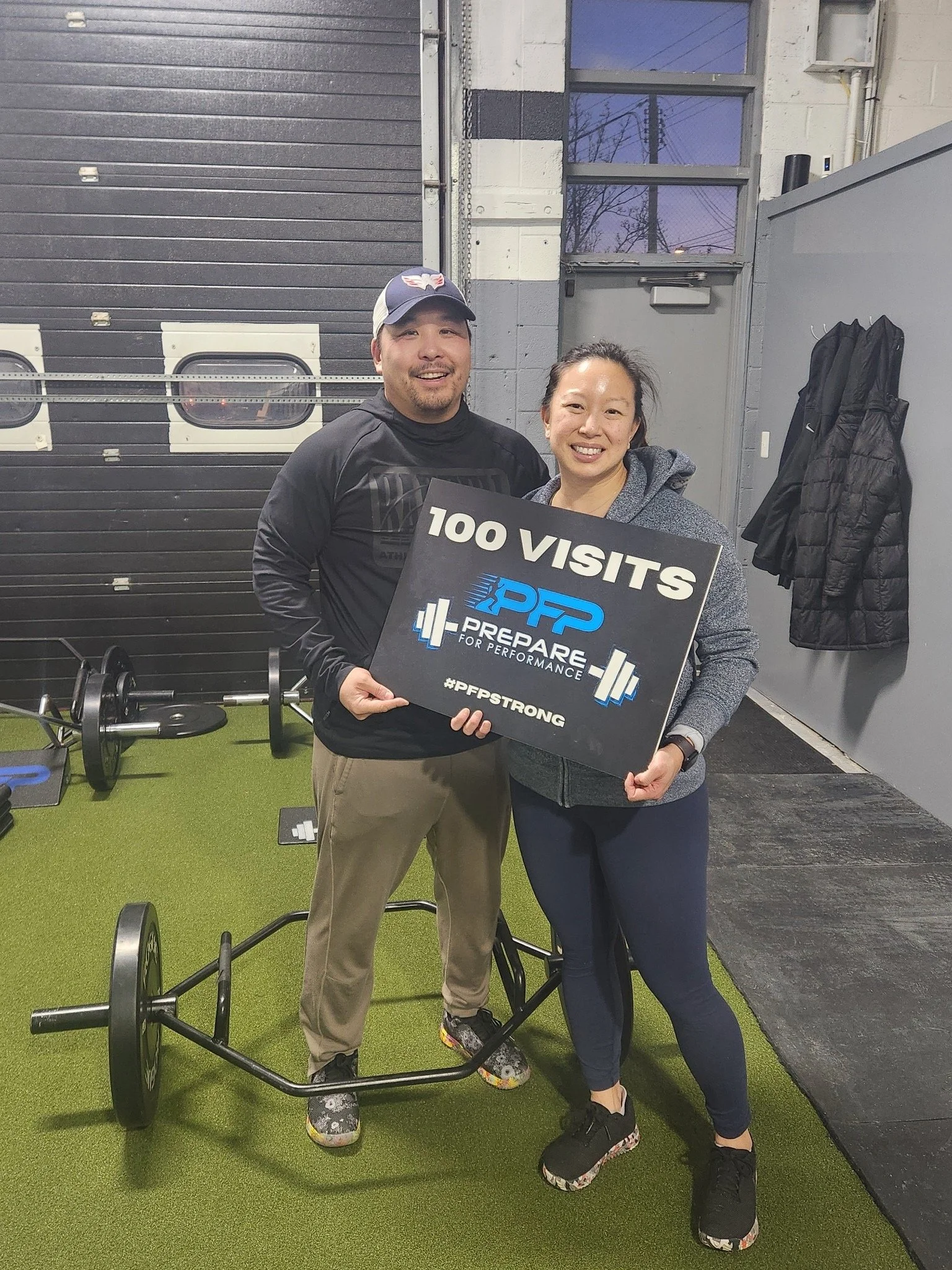 A man and woman smiling indoors, holding a sign that says '100 Visits' with a logo for 'Prepare for Performance' and the hashtag '#PPSTrong'. They are standing on green flooring, with gym equipment in the background.